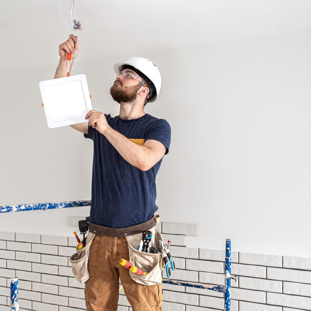 Electrician Builder with beard worker in a white helmet at work, installation of lamps at height. Professional in overalls with a drill on the background of the repair site.